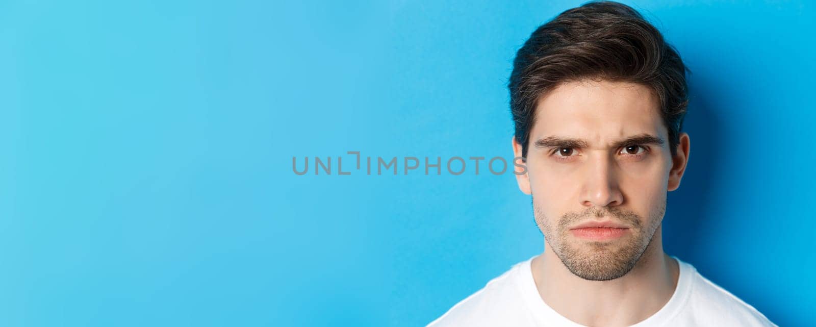 Headshot of angry man frowning, looking disappointed and bothered, standing over blue background.