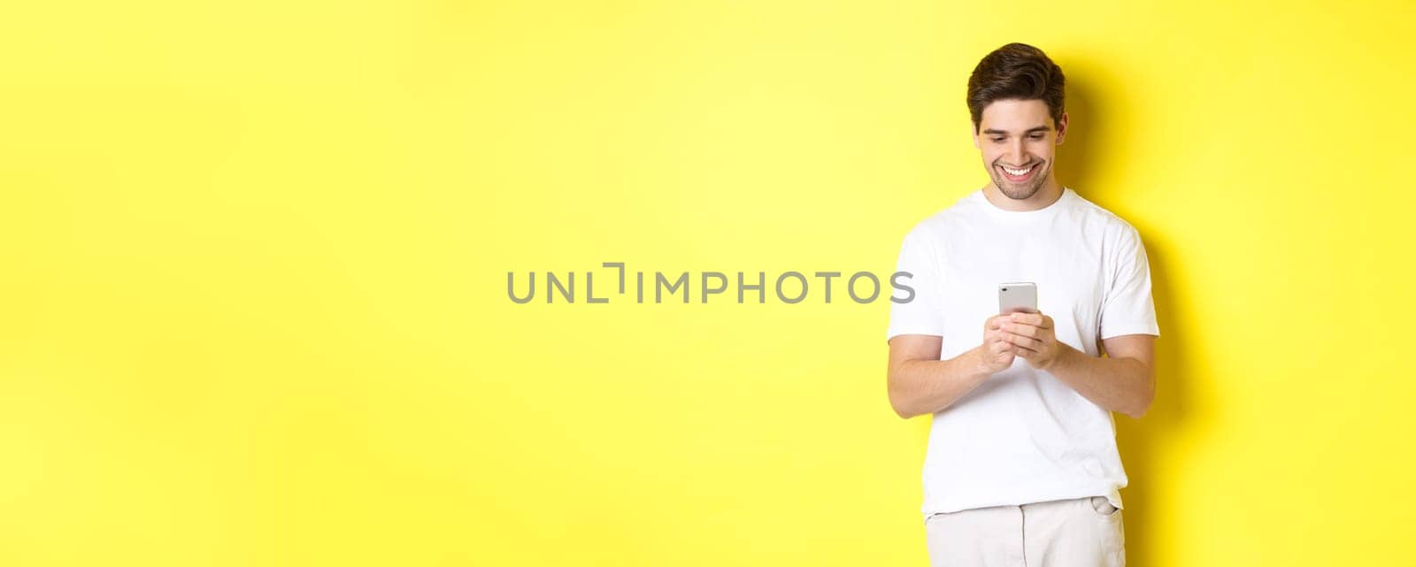 Young man reading text message on smartphone, looking at mobile phone screen and smiling, standing in white t-shirt against yellow background by Benzoix