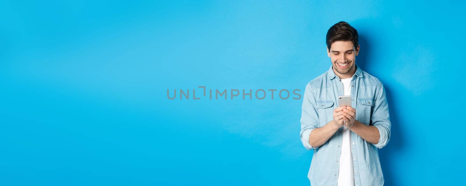 Image of handsome young man using mobile phone, texting on phone and looking pleased, standing over blue background by Benzoix