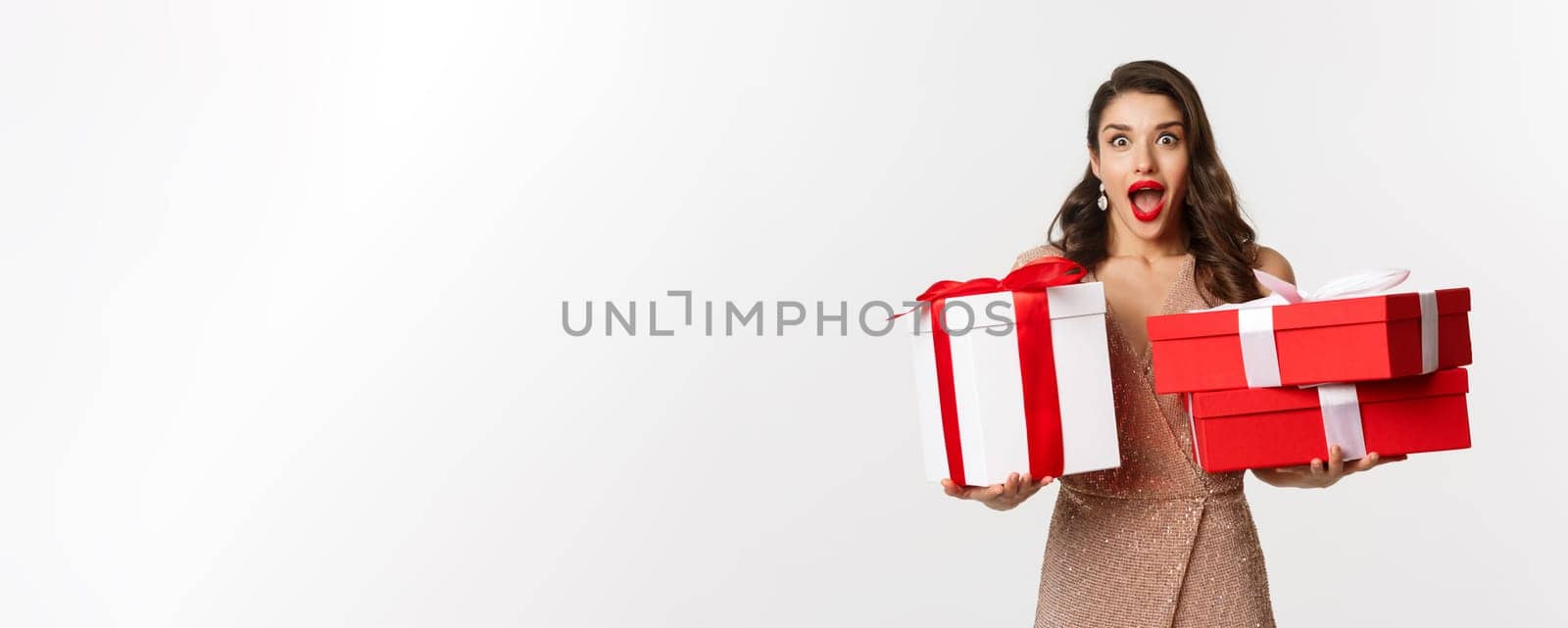 Holidays, celebration concept. Excited and surprised woman holding Christmas gifts and smiling amazed, wearing glamour dress, standing over white background.