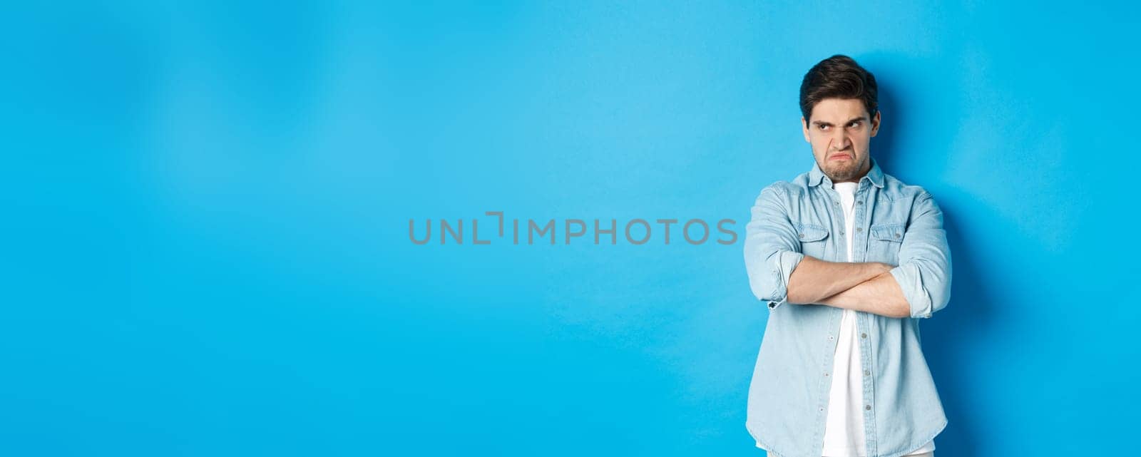 Angry guy cross arms on chest and looking away with insulted expression, standing offended against blue background by Benzoix