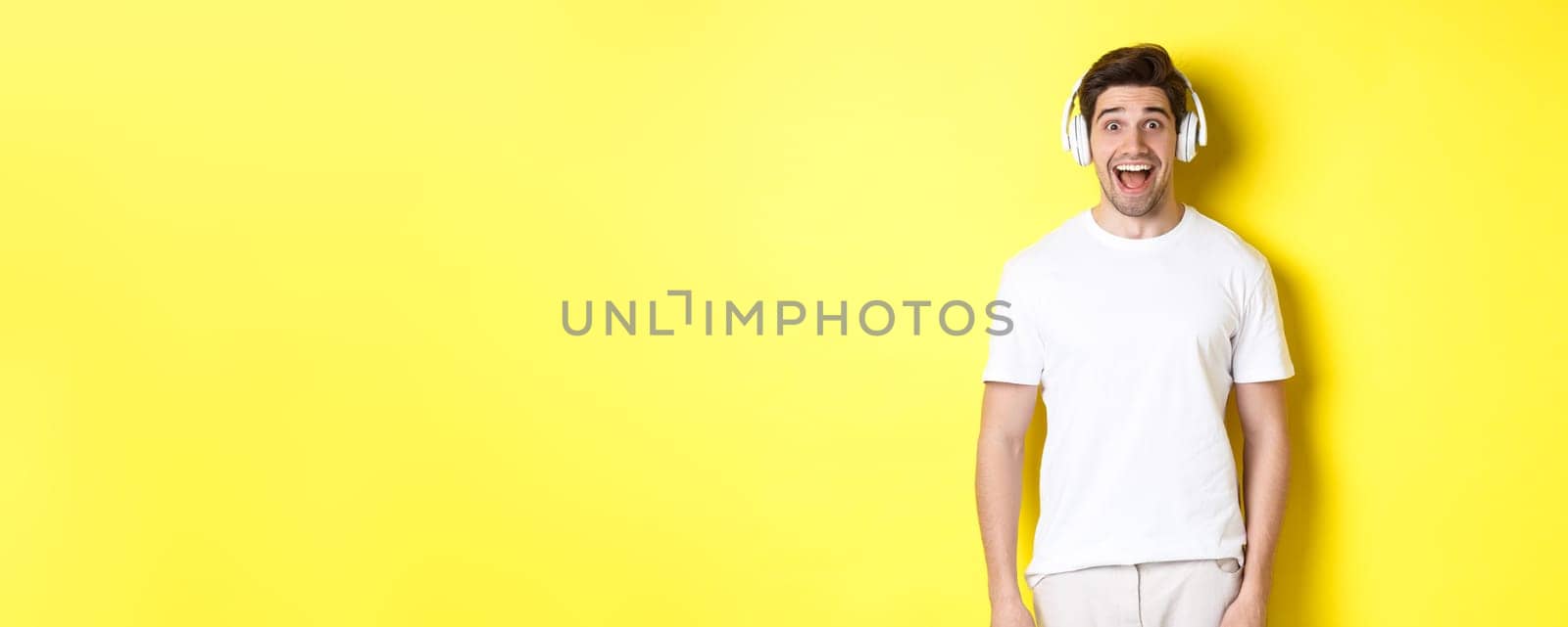 Man in headphones looking surprised, standing against yellow background in white outfit.
