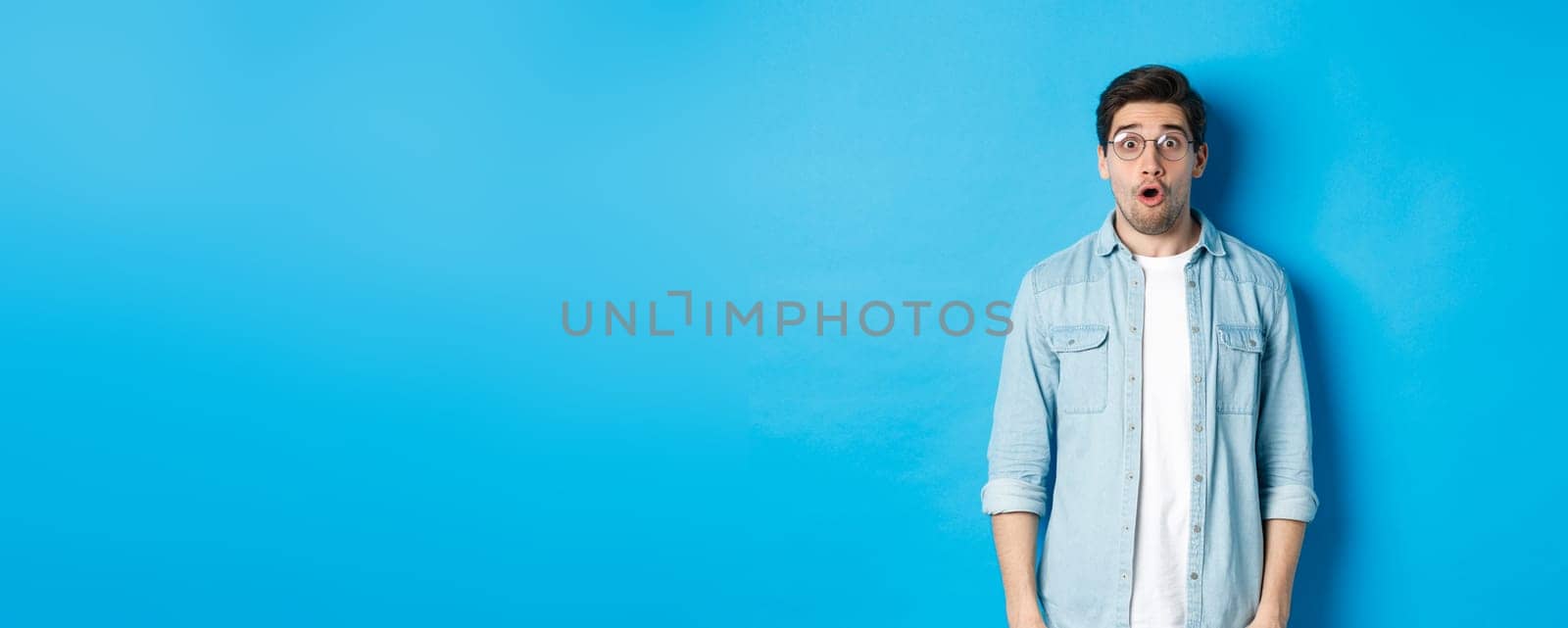 Surprised man in glasses looking at promo offer, saying wow amazed, standing over blue background by Benzoix