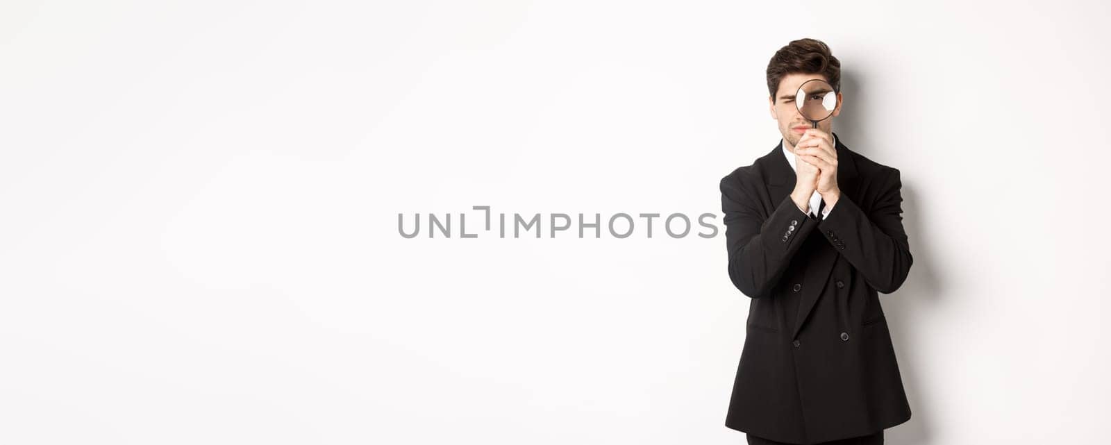 Image of serious businessman in black trendy suit, looking through magnifying glass, looking for employees, standing against white background by Benzoix