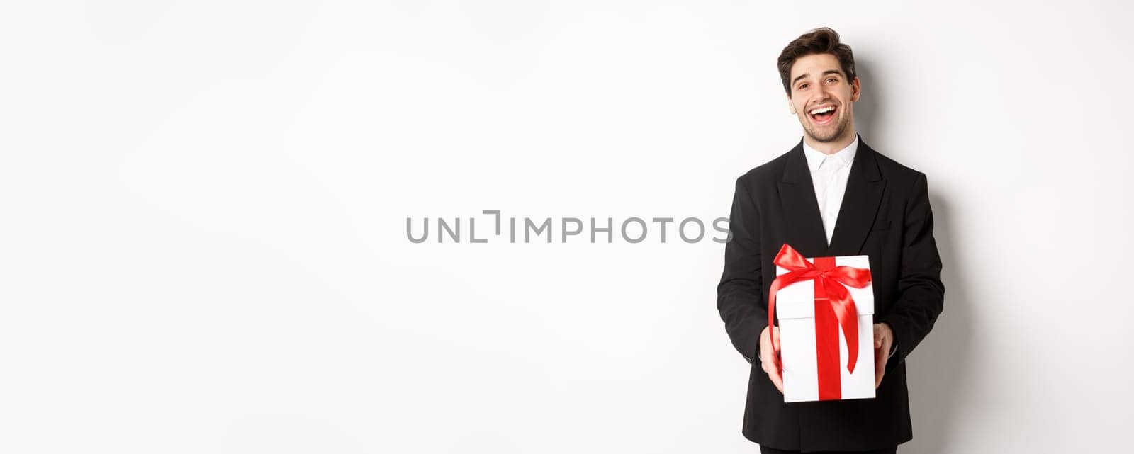 Concept of christmas holidays, celebration and lifestyle. Joyful handsome man in black suit, holding xmas gift and smiling, standing against white background.