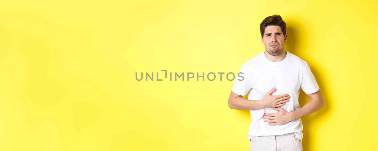 Man having stomach ache, grimacing from pain and touching belly, standing over yellow background by Benzoix