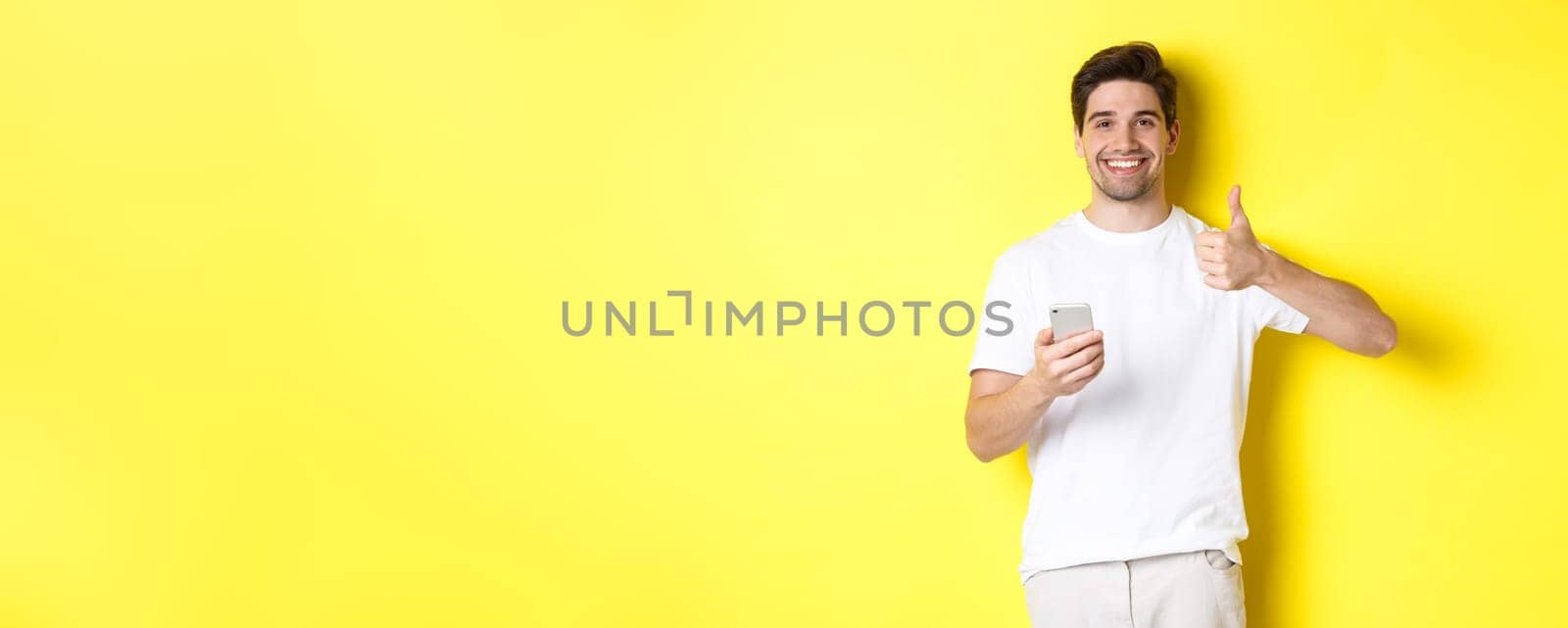 Happy satisfied man holding smartphone, showing thumb up in approval, recommend something online, standing over yellow background.