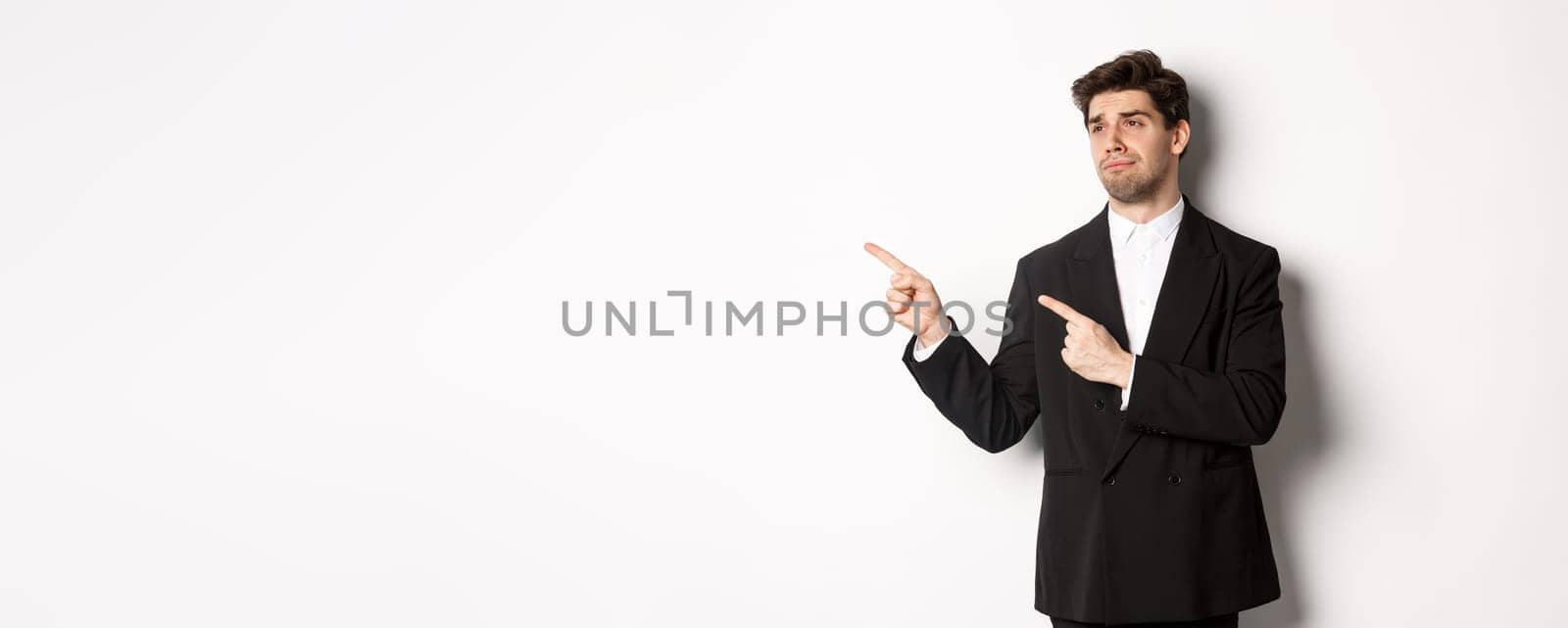 Image of upset and disappointed handsome guy in formal suit, pointing and looking left with sad face, standing over white background.