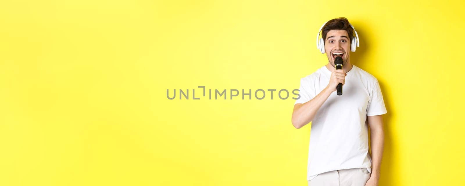 Man in headphones holding microphone, singing karaoke song, standing over yellow background in white clothes.