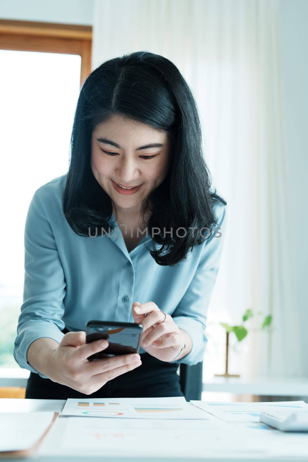 Portrait of a young Asian woman showing a smiling face as she uses her phone, and financial documents on her desk in the early morning hours.