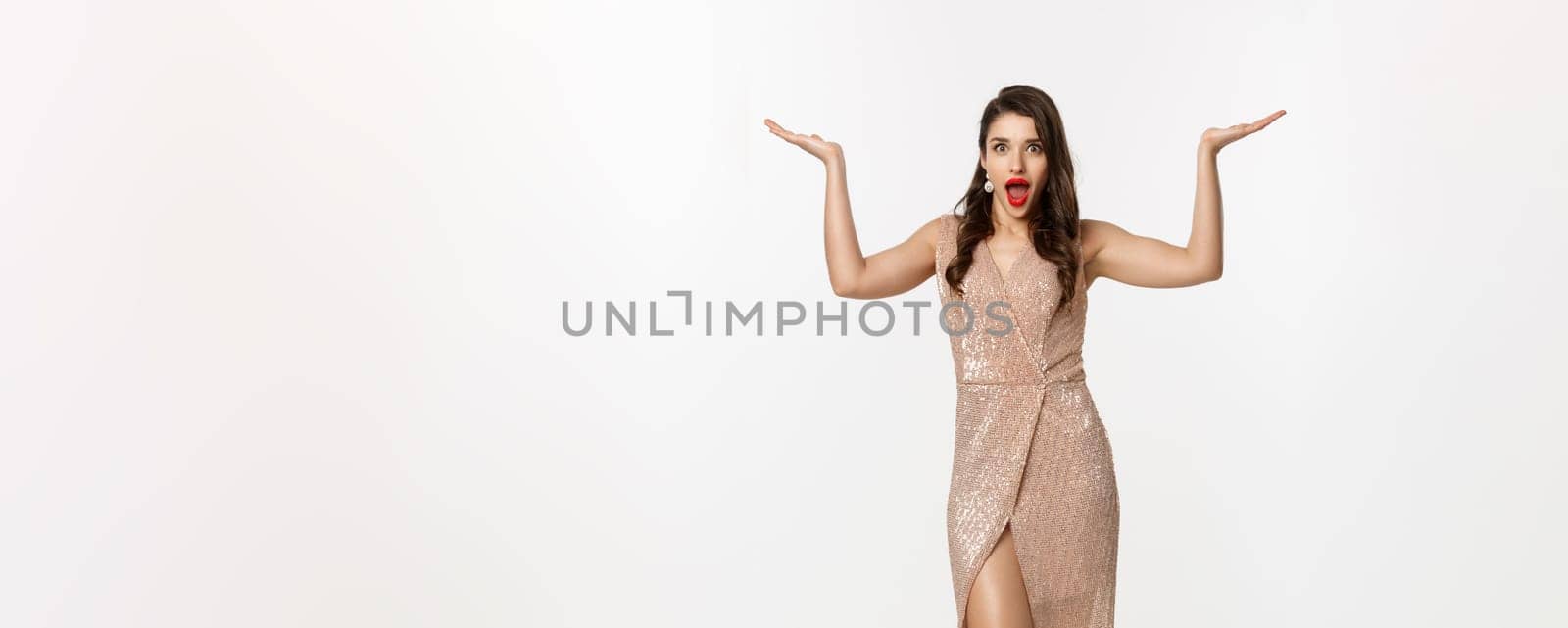 Party and celebration concept. Full-length of beautiful woman in elegant dress, standing near Christmas gifts and looking surprised, standing over white background.