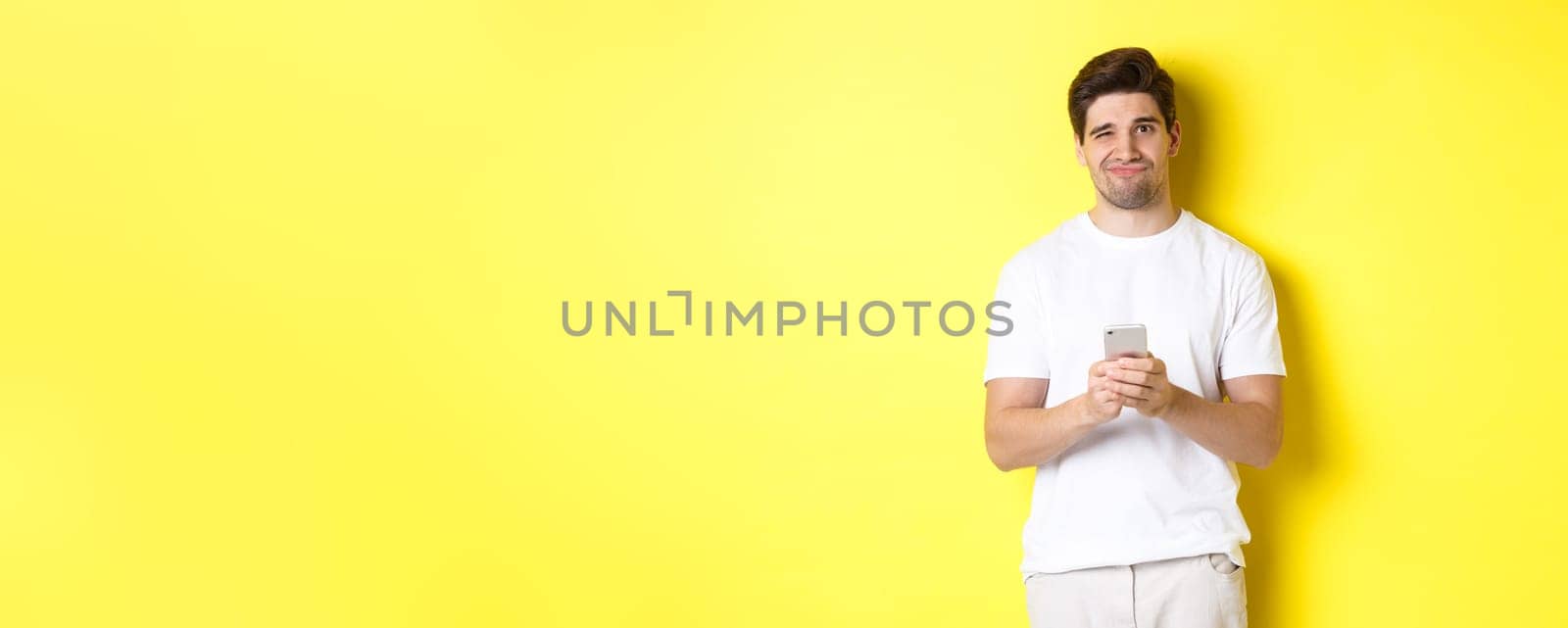Displeased and reluctant man grimacing, being unamused by message on smartphone, standing over yellow background by Benzoix