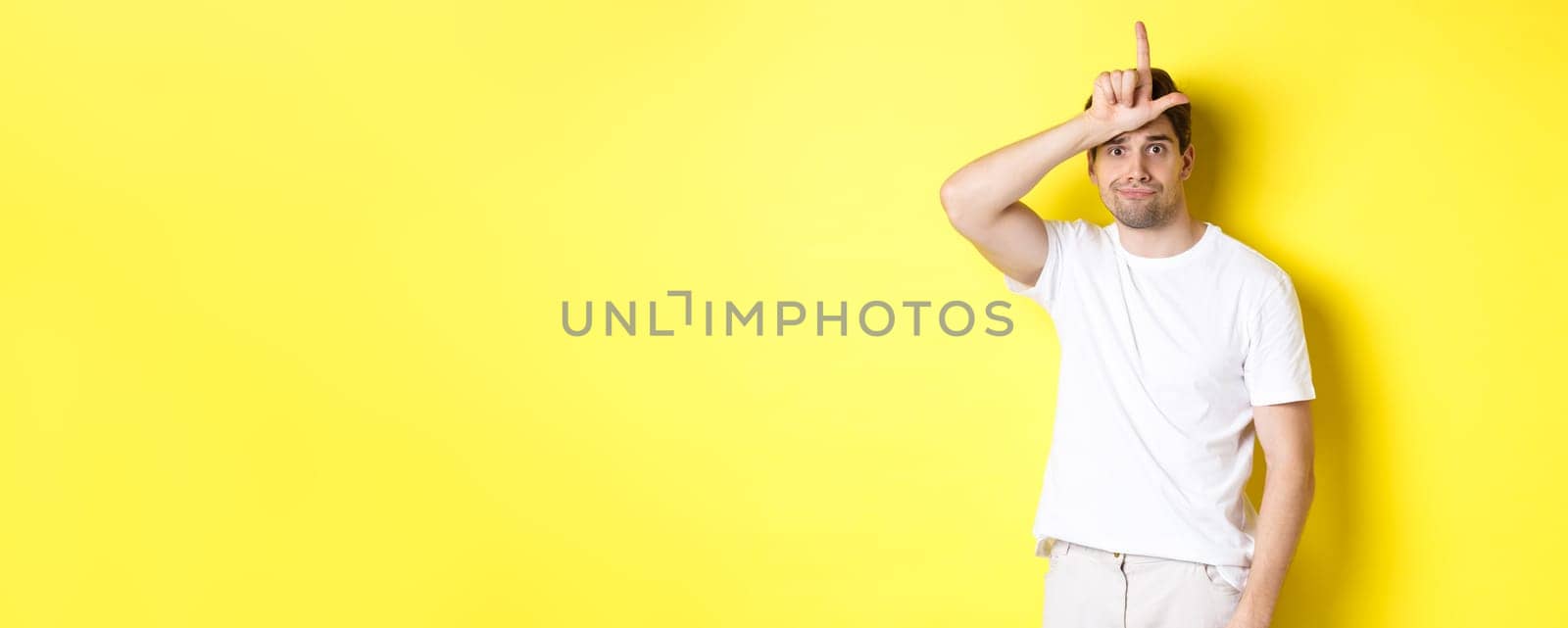 Awkward guy showing loser sign on forehead, looking sad and gloomy, standing in white t-shirt against yellow background.