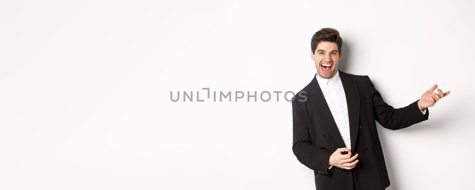 Portrait of happy man dancing at party, playing on invisible guitar and laughing, standing in black suit against white background.