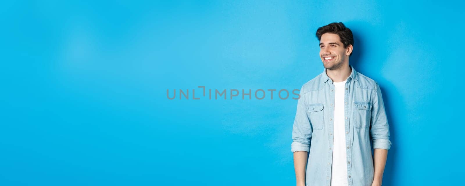 Handsome smiling adult man in casual outfit, smiling and looking left at promo offer, standing against blue background.