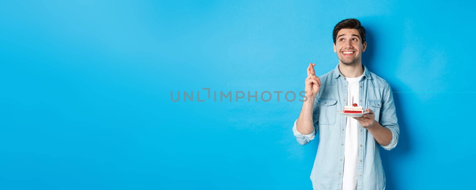 Handsome man making a wish, celebrating birthday, holding b-day cake and cross fingers, standing over blue background.