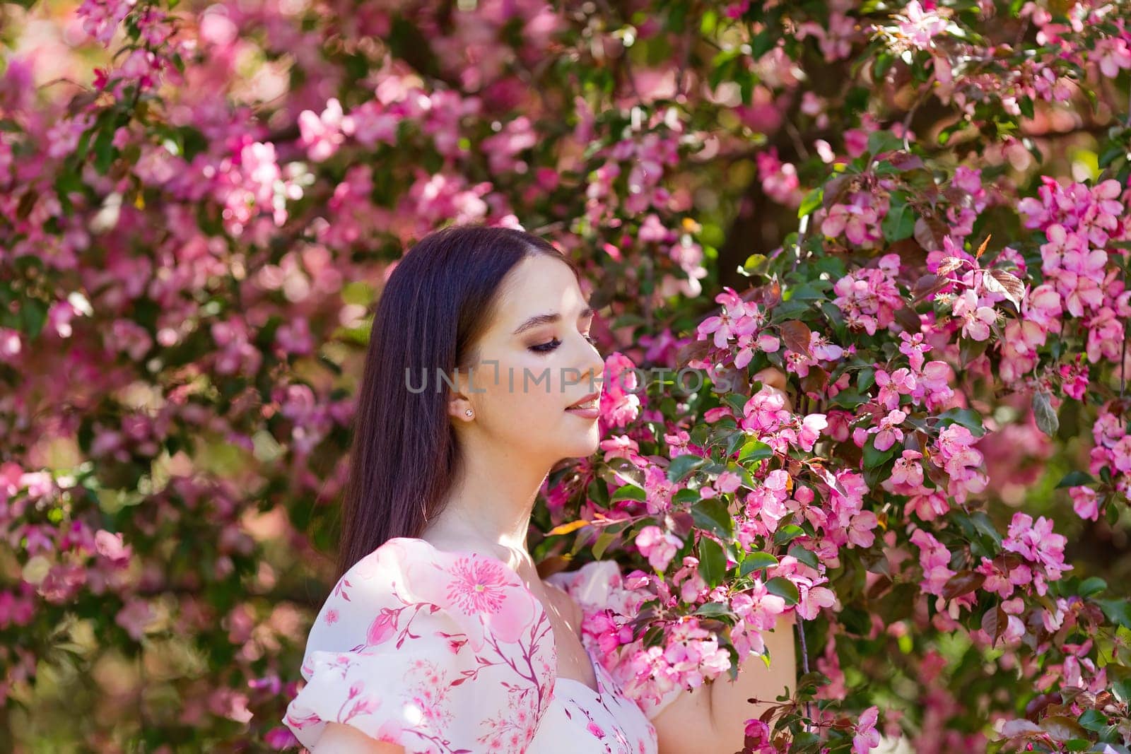 Close up of pretty brunette girl in a pink dress standing under pink blooming apple trees, enjoy, in the spring in the garden. Side view. Close up. Copy space