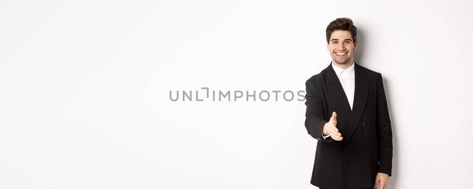 Portrait of handsome businessman in black suit, extending hand for handshake, greet business partners and smiling, welcome to company, standing over white background by Benzoix