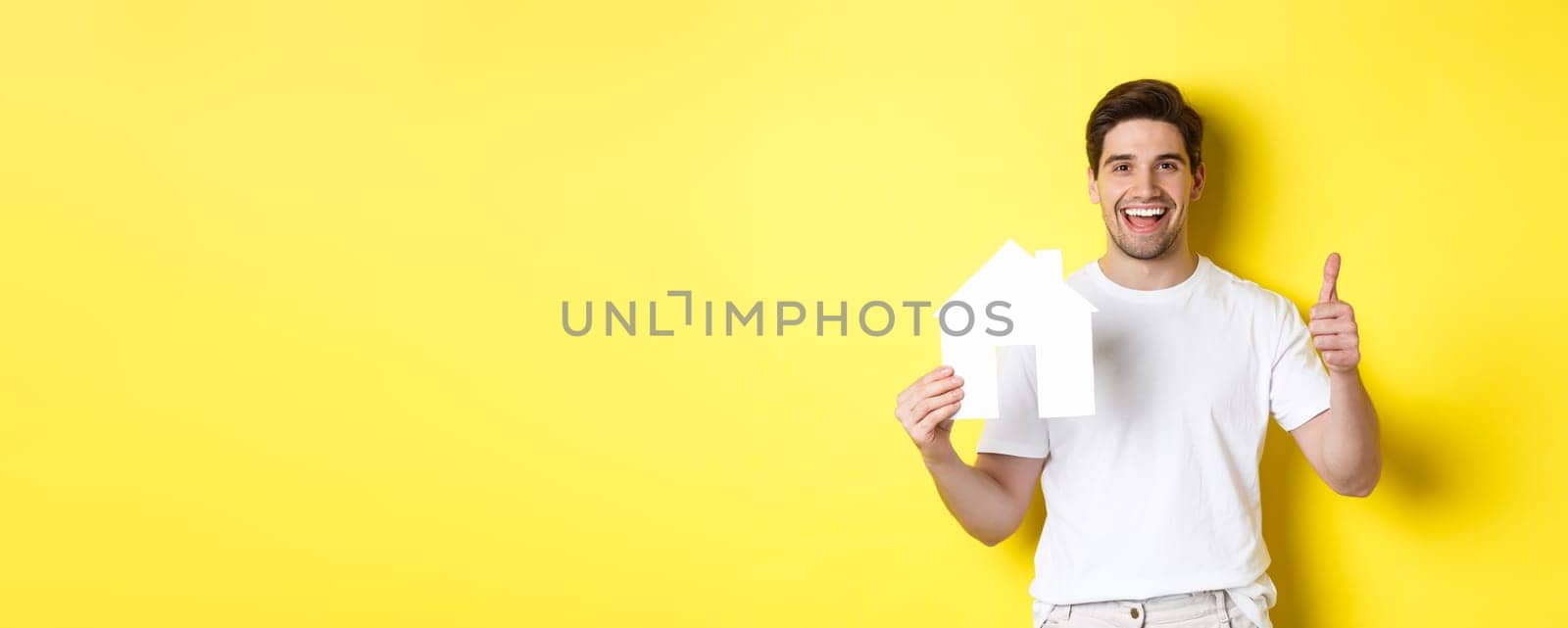 Real estate concept. Happy young man showing paper house model and thumbs up, recommending broker, standing over yellow background.