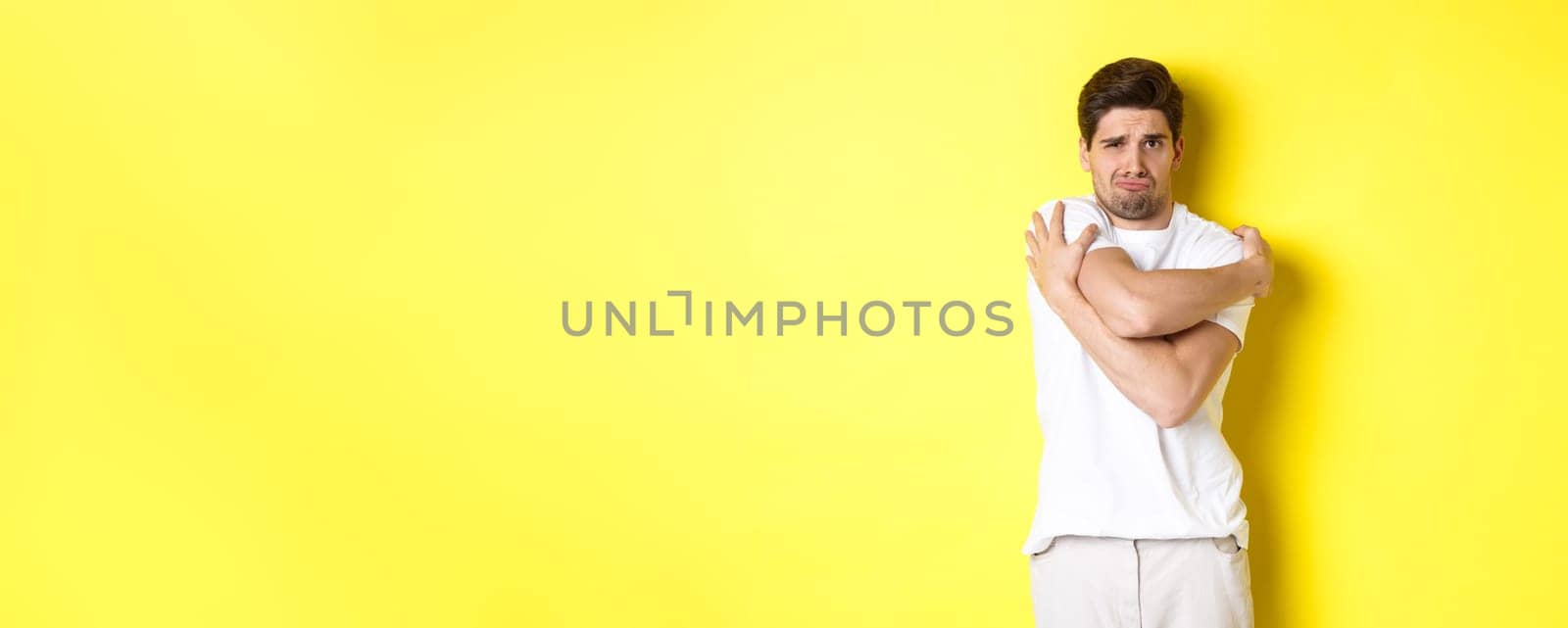 Silly and timid guy trying to comfort himself, hugging his body and frowning scared, standing over yellow background.