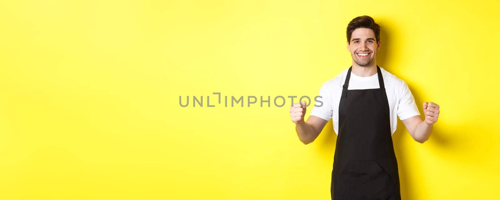 Friendly coffee shop waiter standing with raised hands, place for your sign or logo, standing over yellow background by Benzoix