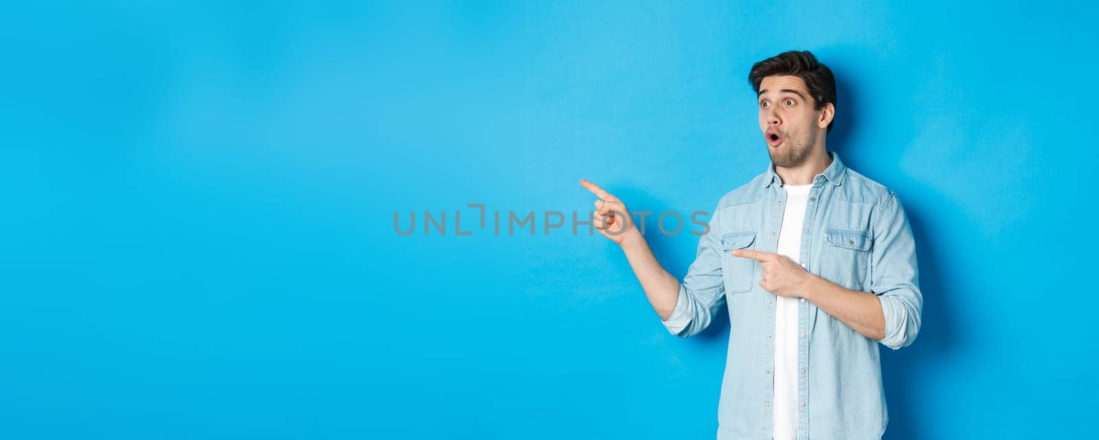 Surprised and amazed man looking at promotion, pointing fingers left at advertisement, standing over blue background.