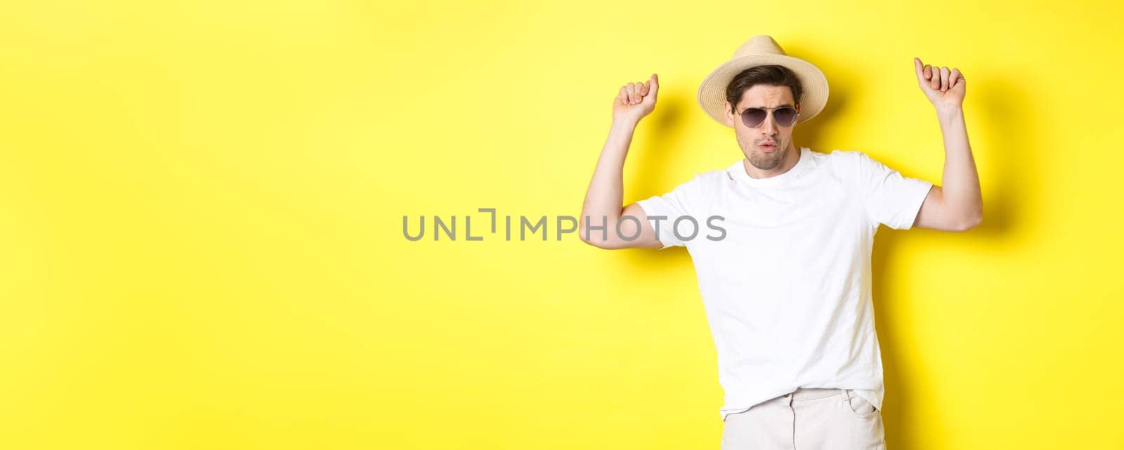Tourism, travelling and holidays concept. Man tourist enjoying vacation, dancing in straw hat and sunglasses, posing against yellow background.