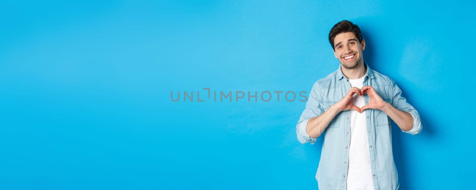 Handsome man smiling, showing heart gesture and looking at camera, saying I love you, standing against blue background.