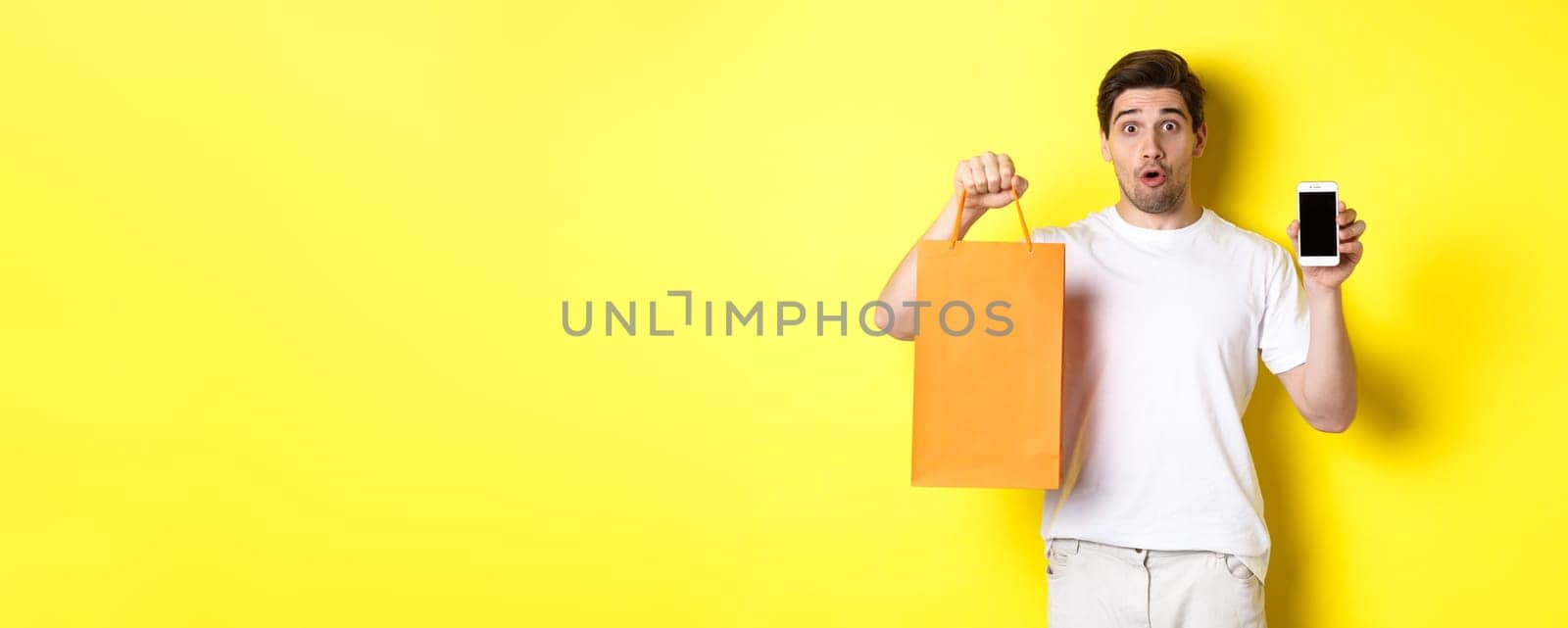 Surprised man showing mobile screen and shopping bag, standing against yellow background. Copy space