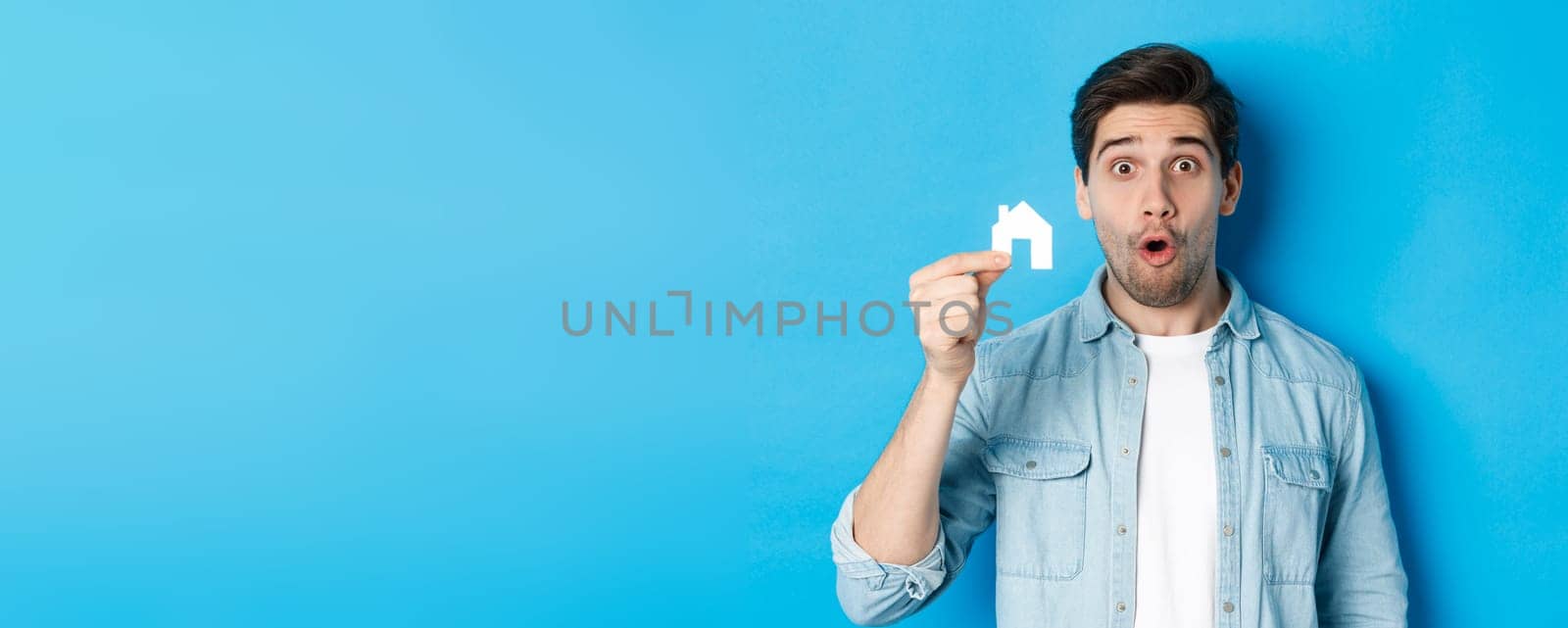 Real estate concept. Surprised guy showing small paper house model, looking amazed, standing against blue background.