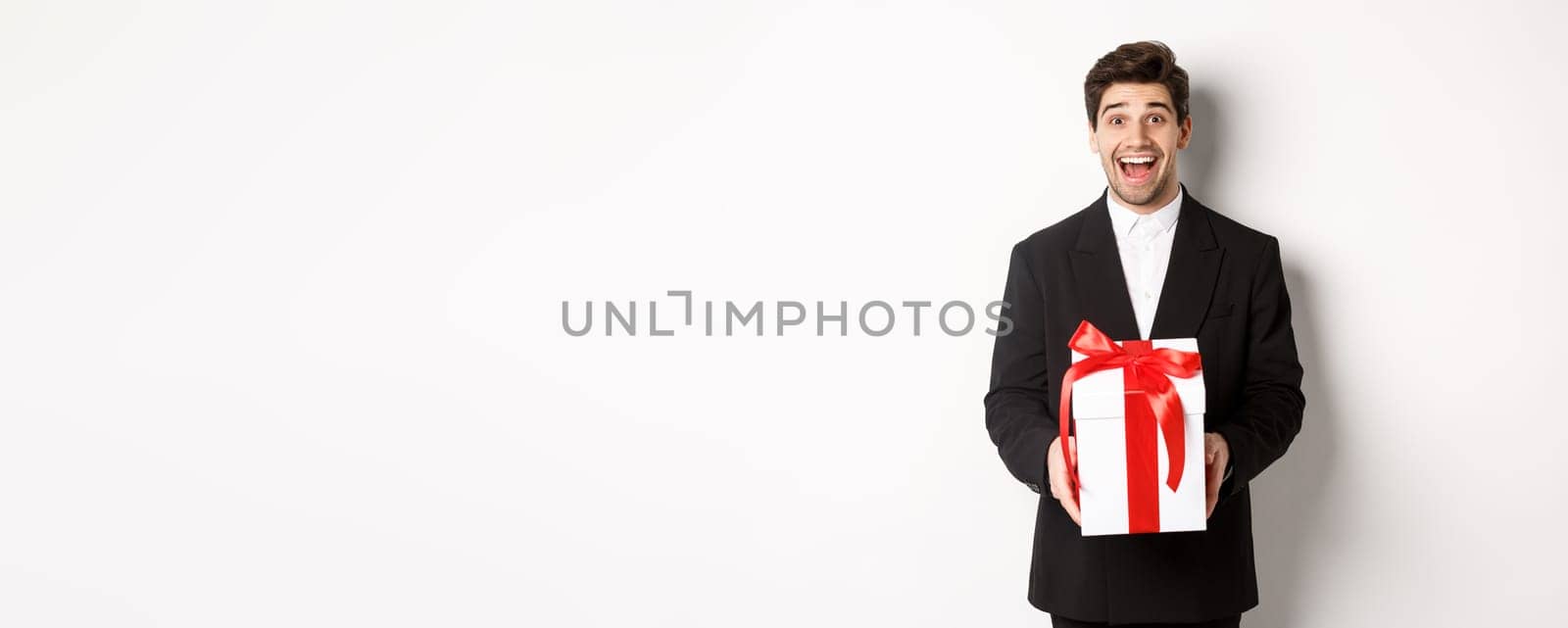 Concept of christmas holidays, celebration and lifestyle. Image of handsome guy in black suit looking excited, have a gift, standing against white background.