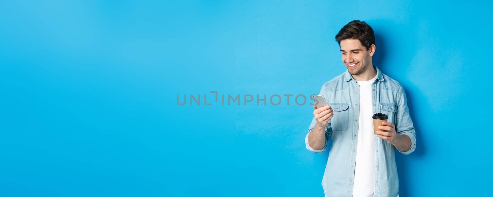 Handsome modern guy reading message on cell phone and drinking coffee, standing against blue background by Benzoix