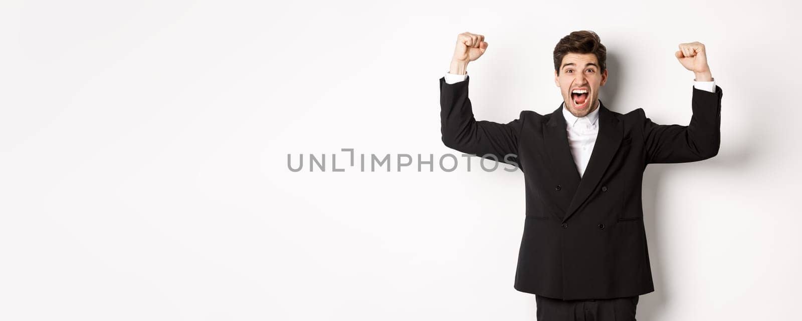 Successful handsome businessman triumphing, raising hands up and shouting yes, rejoicing over achievement, standing against white background.