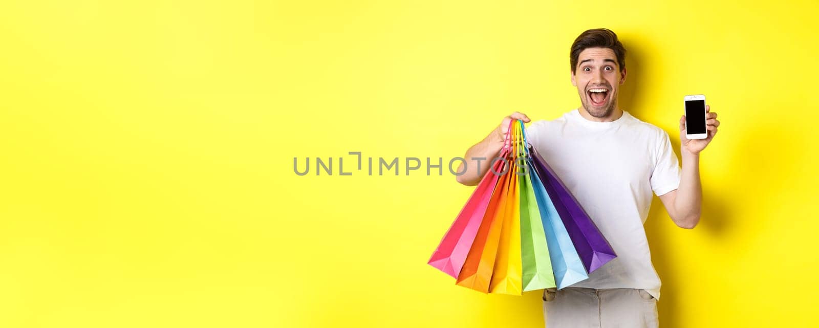 Young man holding shopping bags and showing mobile phone screen, money application, standing over yellow background.