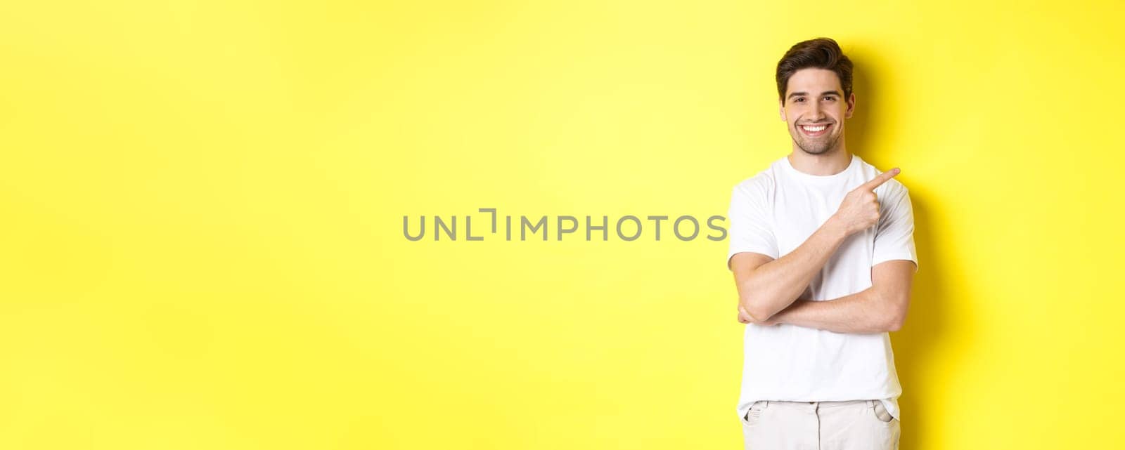 Image of attractive young man pointing finger right at copy space, showing banner or promo offer, standing over yellow background.