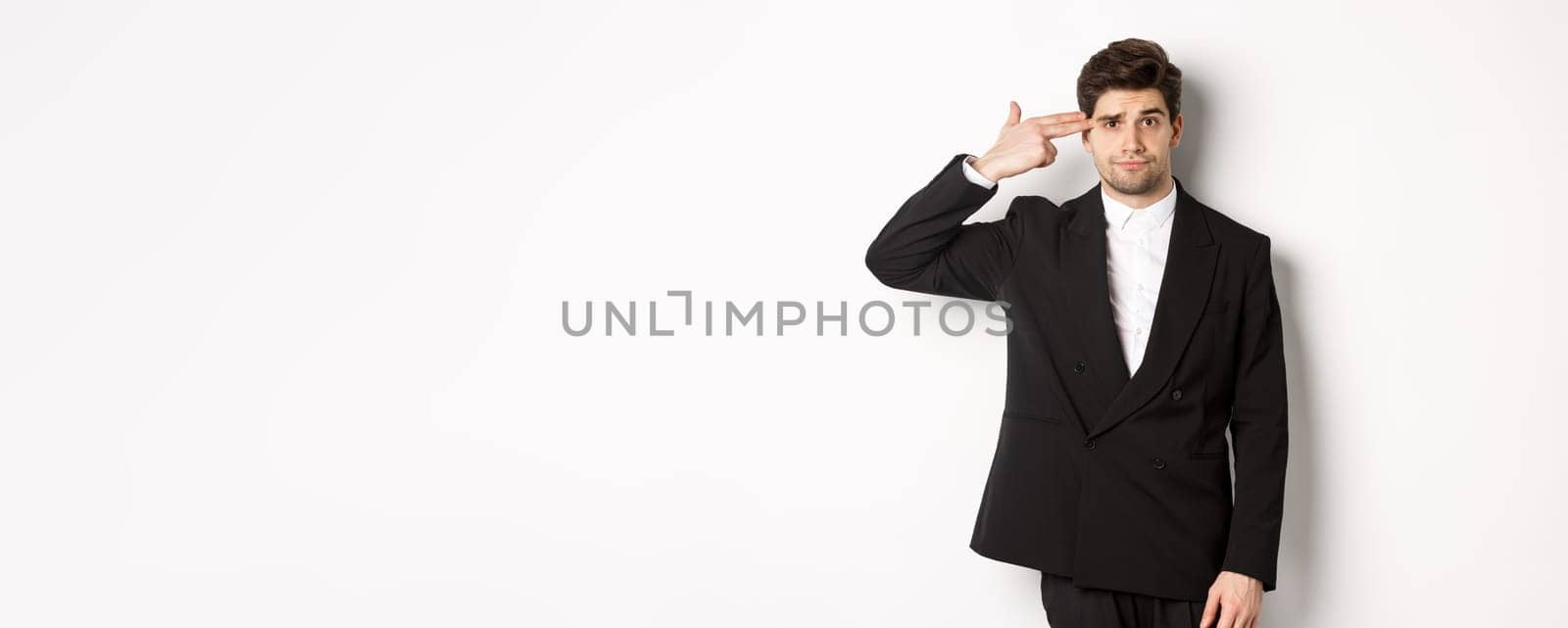 Portrait of distressed and troubled businessman in suit, pointing finger gun at head and looking bothered, shooting himself, standing against white background by Benzoix