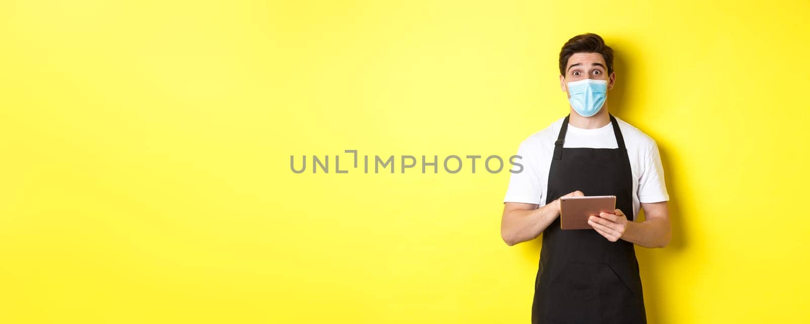 Concept of covid-19, small business and pandemic. Waiter in black apron and medical mask taking order, holding digital tablet, standing over yellow background by Benzoix