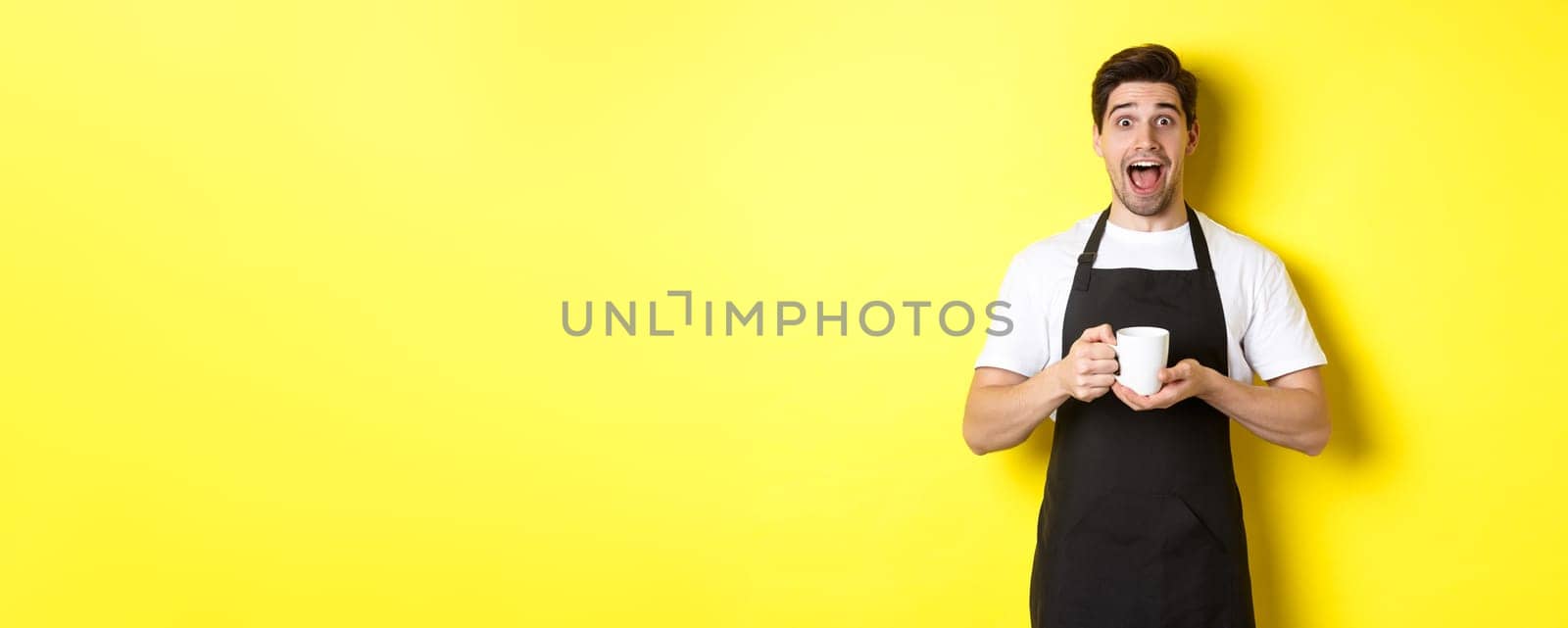 Young male barista holding coffee cup and looking surprised, standing in black apron against yellow background by Benzoix