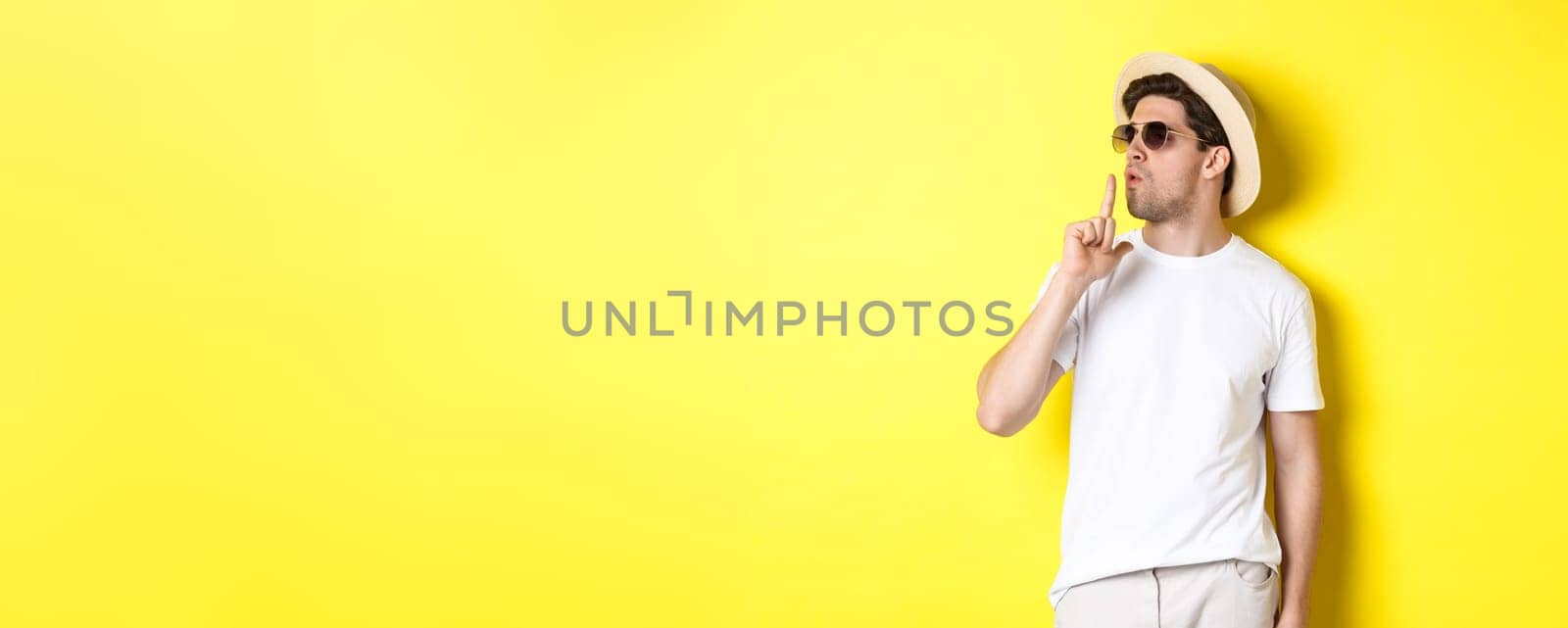 Cool young male tourist blowing at finger gun and looking confident, standing against yellow background. Vacation and lifestyle concept.