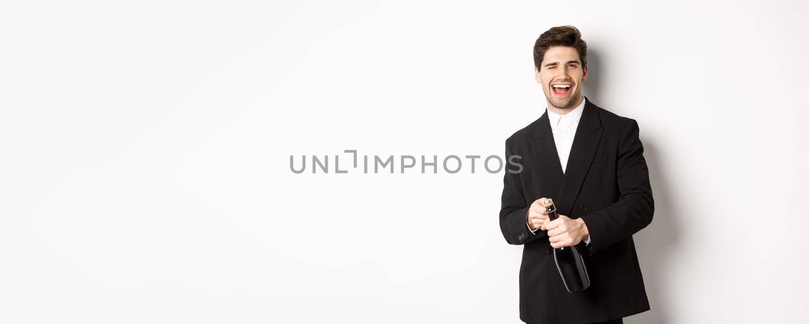 Image of handsome, confident man in black suit, celebrating holiday, open a bottle of champagne and partying, standing joyful against white background by Benzoix