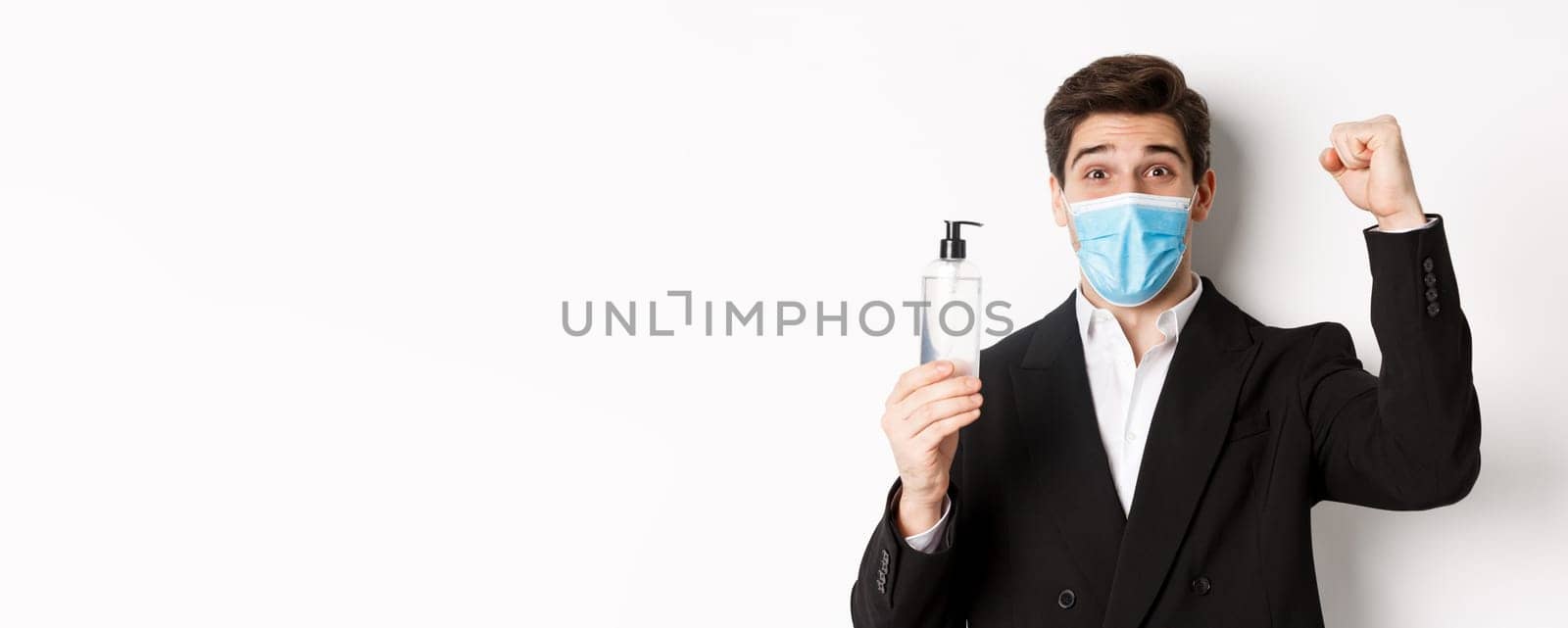 Concept of covid-19, business and social distancing. Close-up of happy man in trendy suit and medical mask, cheering and raising hand up, showing hand sanitizer, white background.