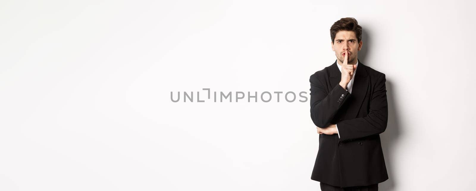 Portrait of angry boss in suit shushing at you, telling to be quiet, showing taboo hush sign and frowning, standing over white background.