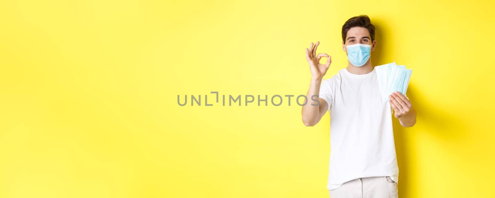 Concept of covid-19, quarantine and preventive measures. Satisfied man showing okay sign and giving medical masks, standing over yellow background.