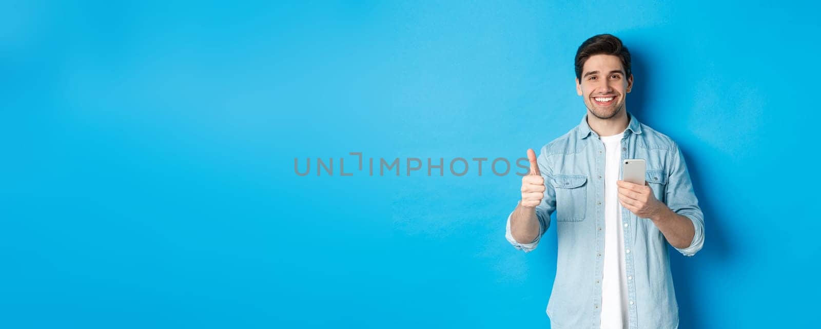 Concept of online shopping, applications and technology. Satisfied man in casual clothes smiling, showing thumbs up after using smartphone app, standing over blue background.