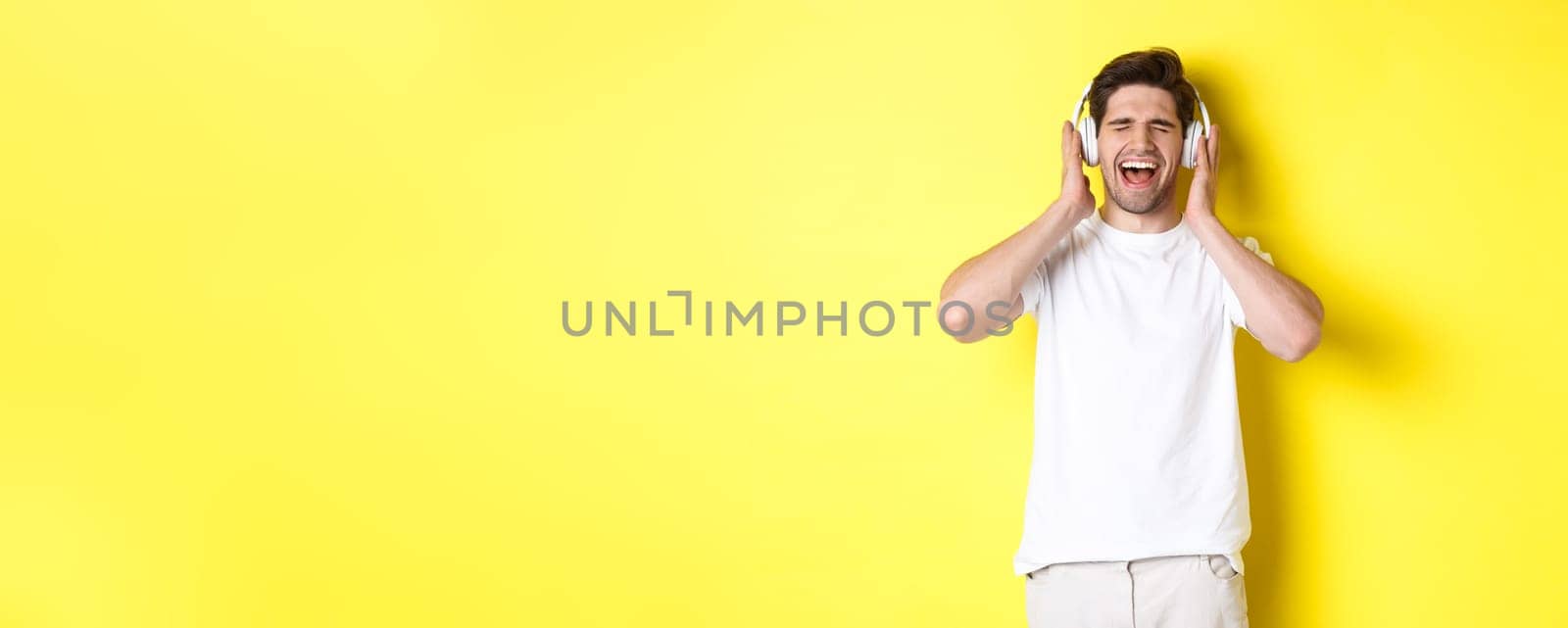 Happy guy listening music in new headphones, buying earphones on black friday, standing over yellow background by Benzoix