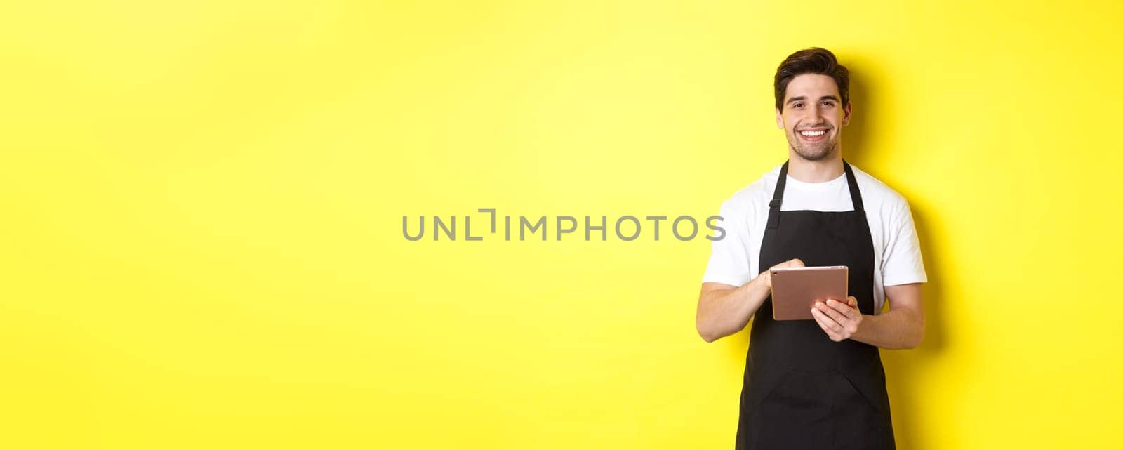 Handsome waiter taking orders, holding digital tablet and smiling, wearing black apron uniform, standing over yellow background.