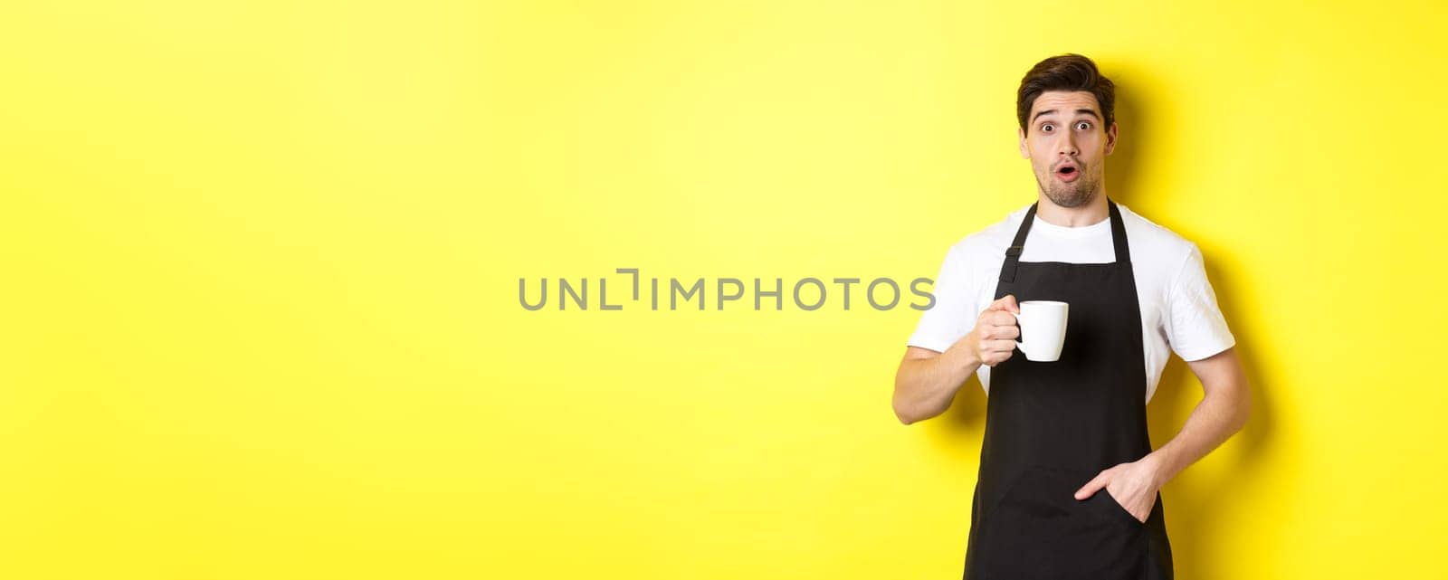 Barista holding coffee mug and looking surprised, standing in black apron cafe uniform against yellow background by Benzoix
