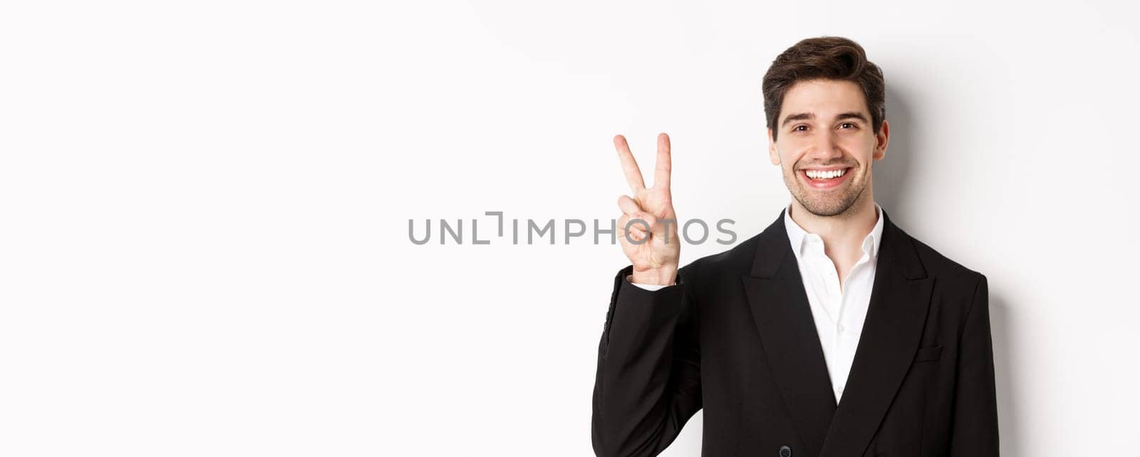 Close-up of handsome businessman in black suit, smiling amazed, showing number two, standing over white background by Benzoix