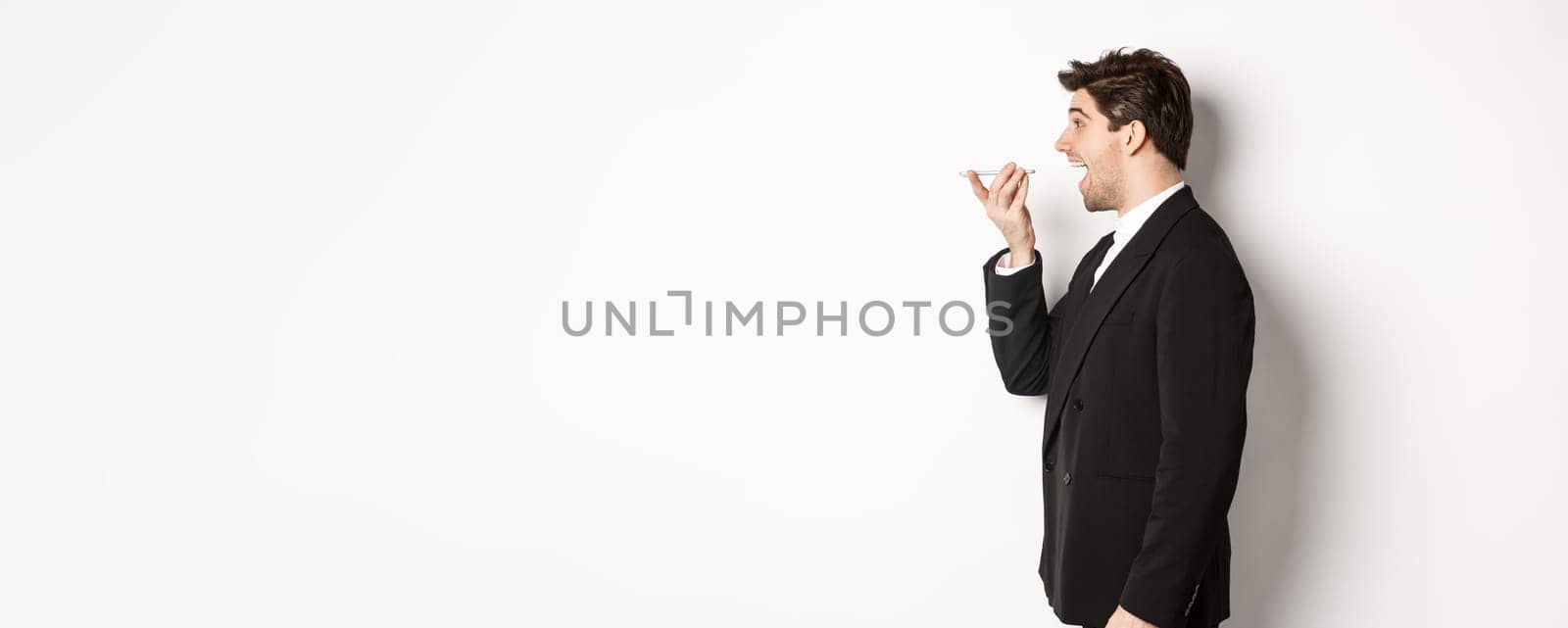 Profile shot of handsome businessman in black suit talking on speakerphone, smiling and looking happy, recording voice message, standing over white background by Benzoix