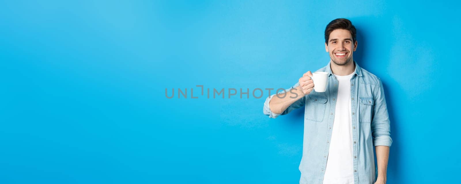 Smiling bearded man holding mug and drinking coffee, standing against blue background.